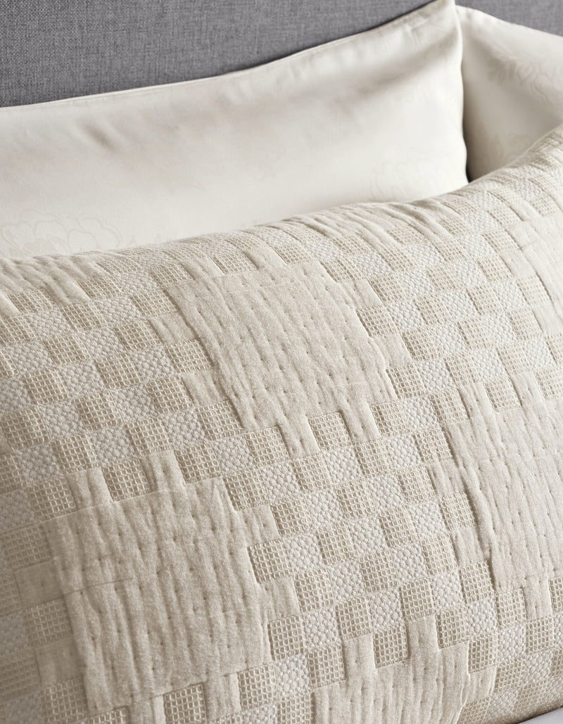 A close-up of two Cozy Earth Sonoma Stitch Shams in off-white, one showcasing a subtle floral pattern and the other displaying a textured, geometric woven design, arranged on a grey upholstered surface.