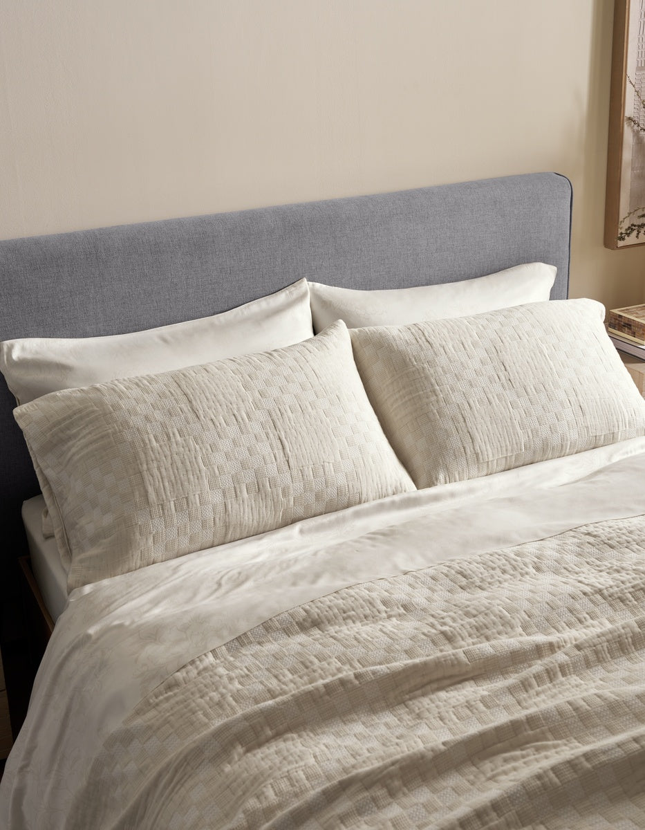 A neatly made bed featuring Sonoma Stitch Shams by Cozy Earth, a gray upholstered headboard, cream-colored pillows, and a textured cream blanket in a softly lit bedroom.