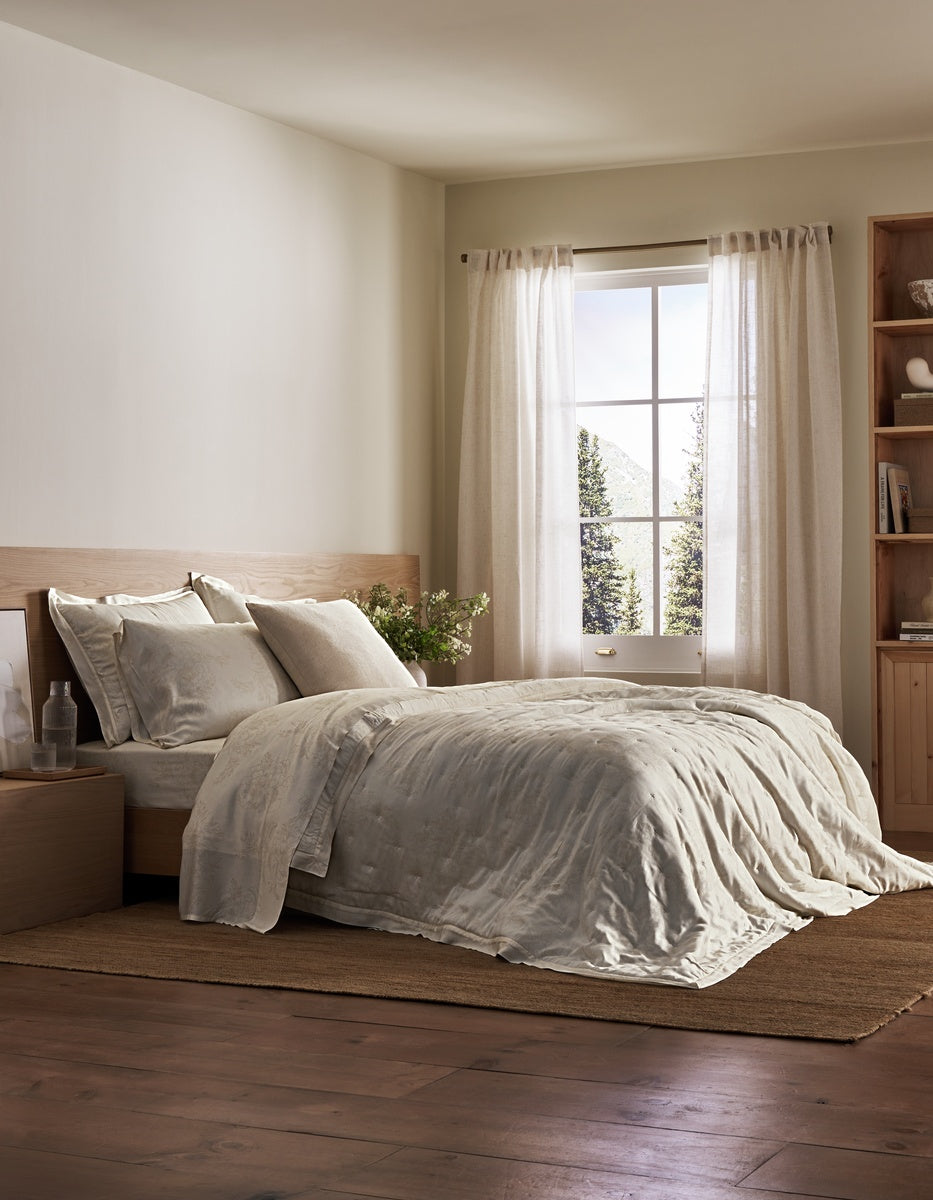 A cozy bedroom features a neatly made bed adorned with the Sonoma Throw Pillow from Cozy Earth, light bedding, a wooden nightstand, and a bookcase. Sunlight streams through sheer curtains on the window, illuminating the room’s neutral tones and hardwood floor.