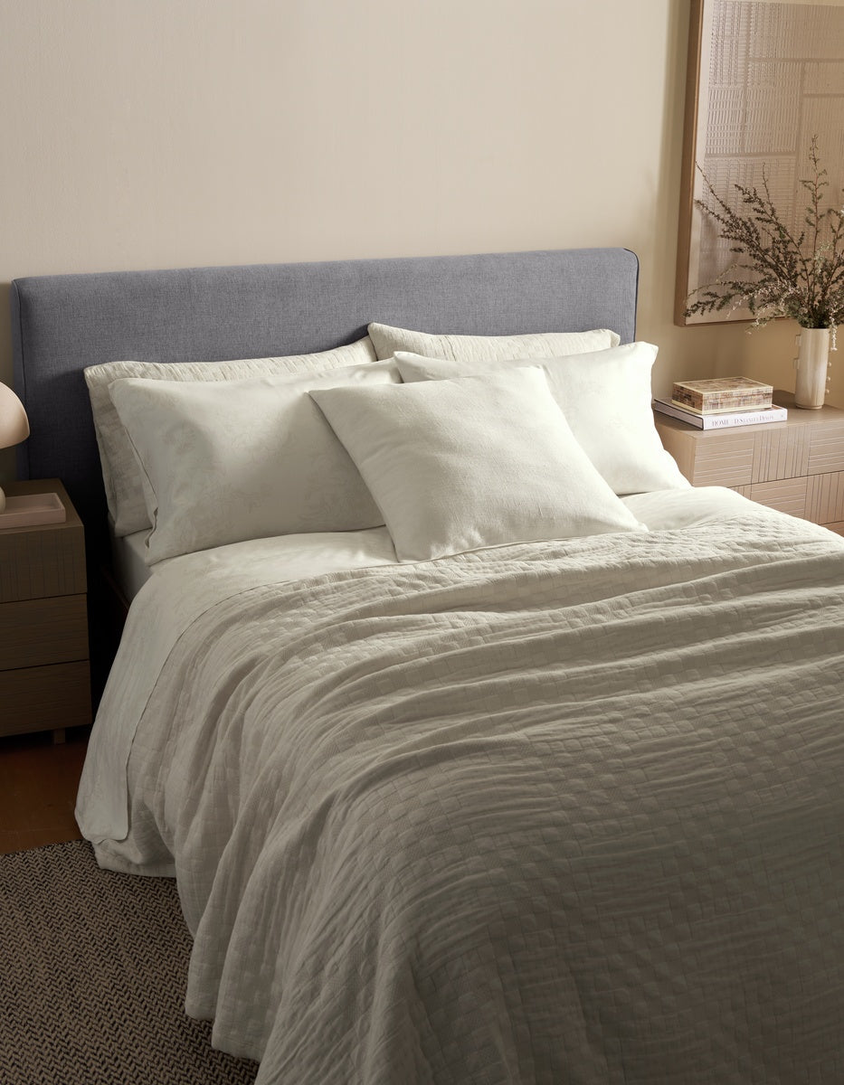 A modern bedroom with a neatly made bed featuring white bedding and pillows, including the Sonoma Throw Pillow by Cozy Earth, a gray upholstered headboard, and light wood nightstands with a vase of greenery.