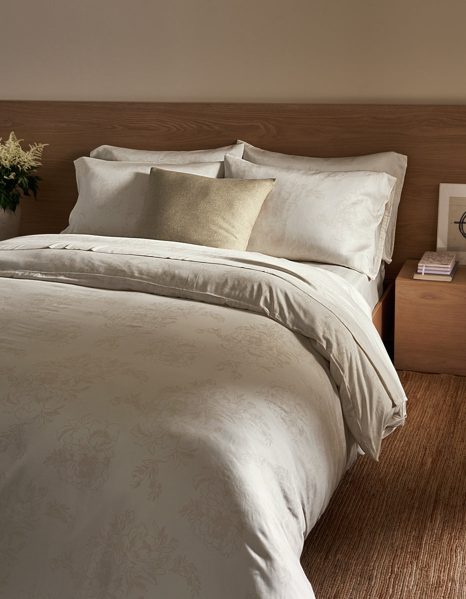 A neatly made bed with cream-colored bedding and a single Cozy Earth Sonoma Throw Pillow in beige, next to a wooden nightstand with stacked books and a small flower arrangement.