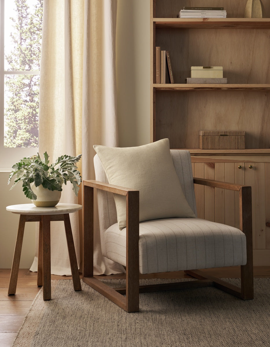 A cozy reading nook with a wooden armchair, a Cozy Earth Sonoma Throw Pillow, a round side table topped with a potted plant, and built-in wooden shelves in a softly lit room.