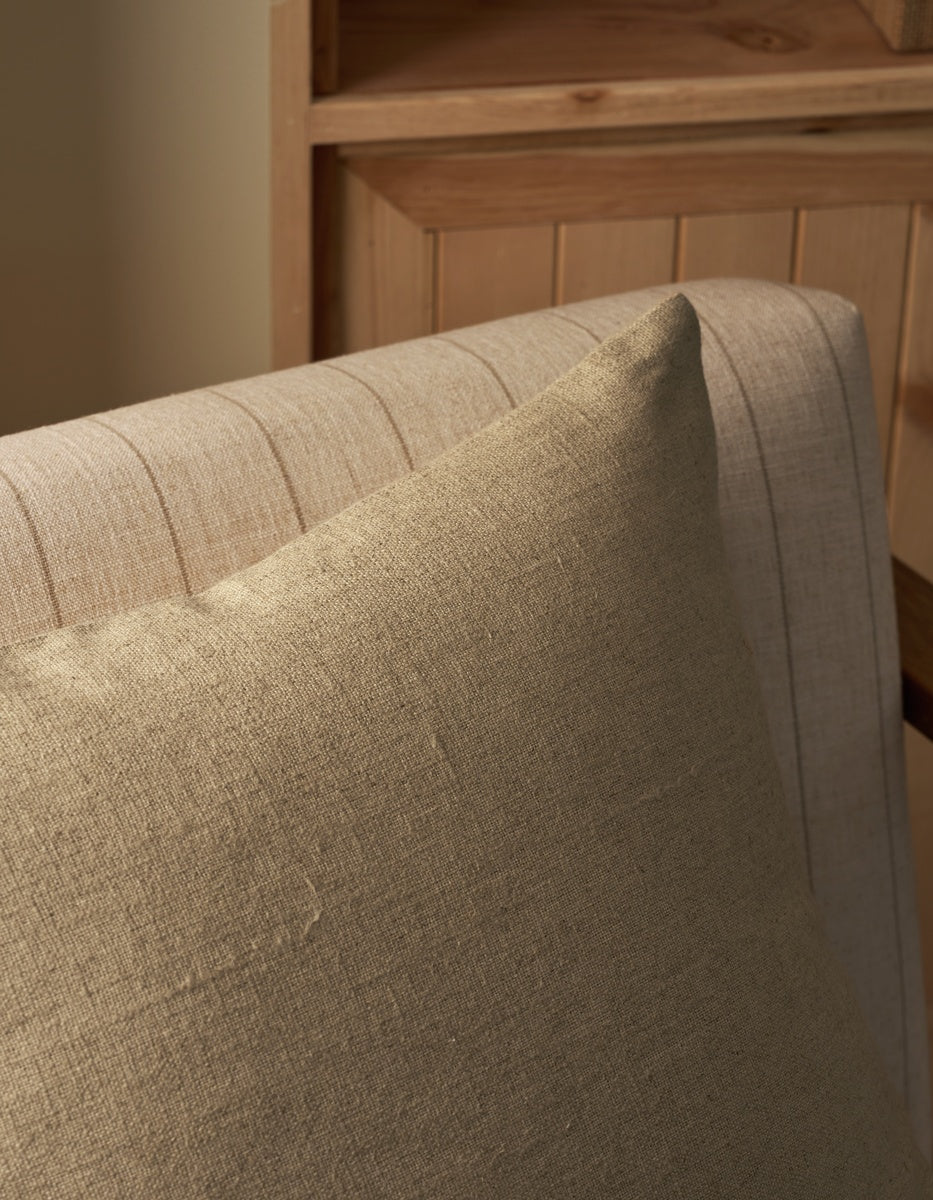 A close-up of the Sonoma Throw Pillow by Cozy Earth, in beige, resting on a light-colored striped upholstered chair, with wooden furniture in the background.