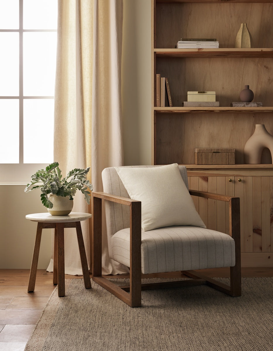 A cozy reading nook featuring a wooden armchair topped with the Cozy Earth Sonoma Throw Pillow, a round side table holding a potted plant, built-in shelves, and a window adorned with light curtains that let in soft natural light.