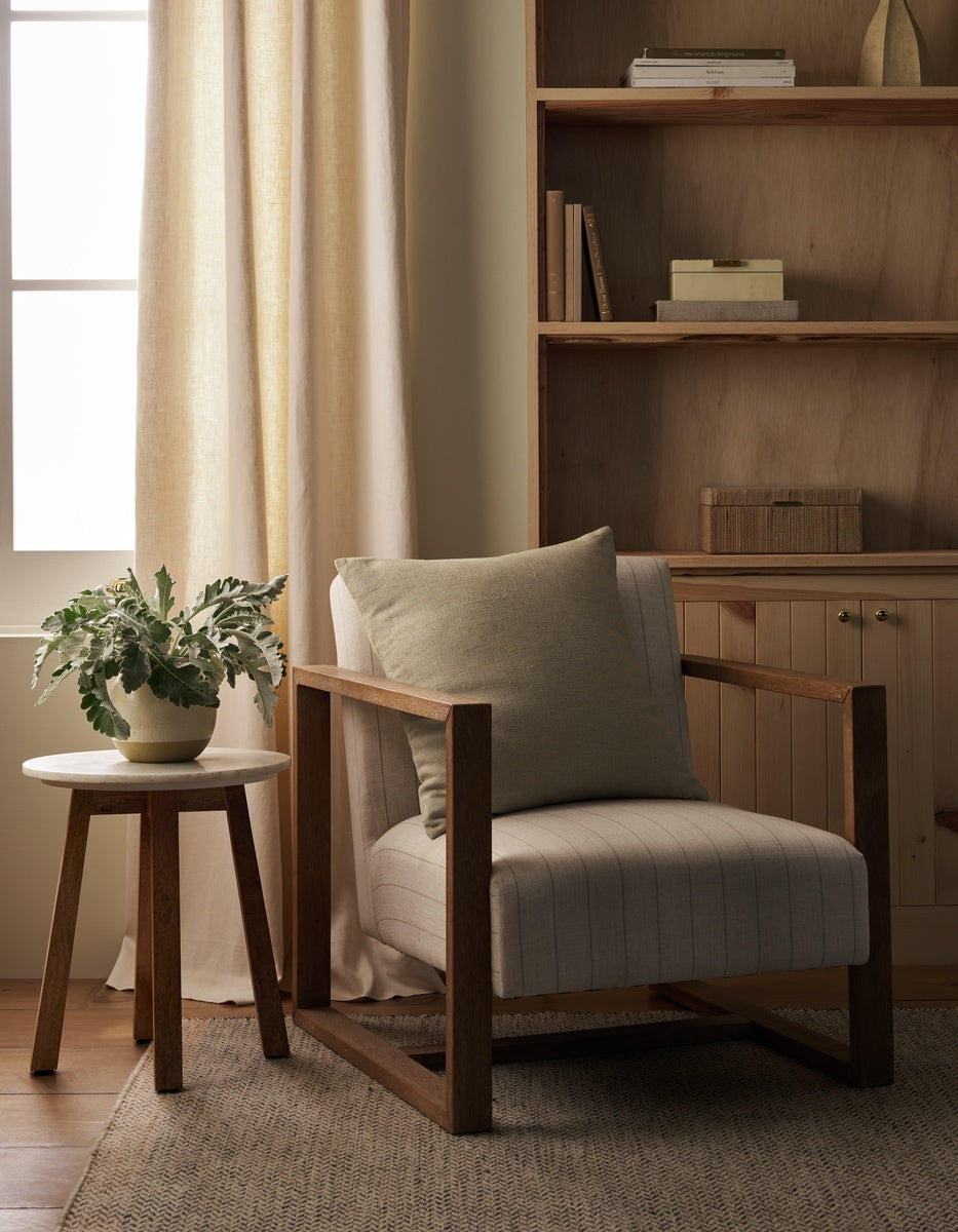 A cozy reading nook with a striped armchair, a neutral Sonoma Throw Pillow by Cozy Earth, a wooden side table holding a potted plant, built-in wooden shelves, a closed cabinet, and sunlight streaming through a window with light curtains.|Color:Tallow |Size:26" x 26"