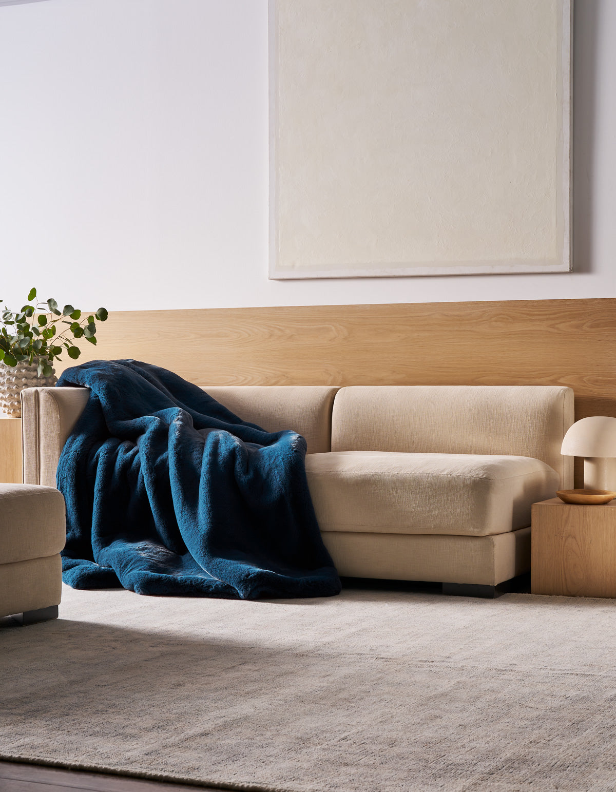 A beige sectional sofa is draped with the Cozy Earth Superplush Cuddle Blanket in deep blue, positioned beside a small wooden side table topped with a lamp and a plant. The space features a light rug, wood paneling, and a large blank canvas displayed on the wall.
