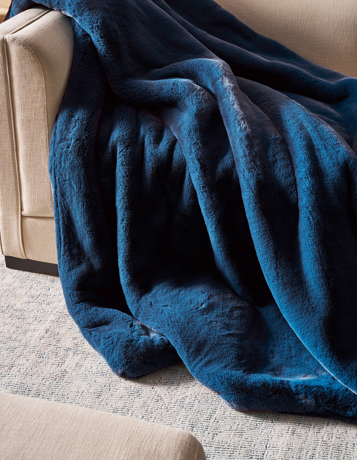A Superplush Cuddle Blanket by Cozy Earth in rich dark blue is draped over the arm and seat of a beige sofa, with part of the blanket pooling onto a light-colored textured rug.