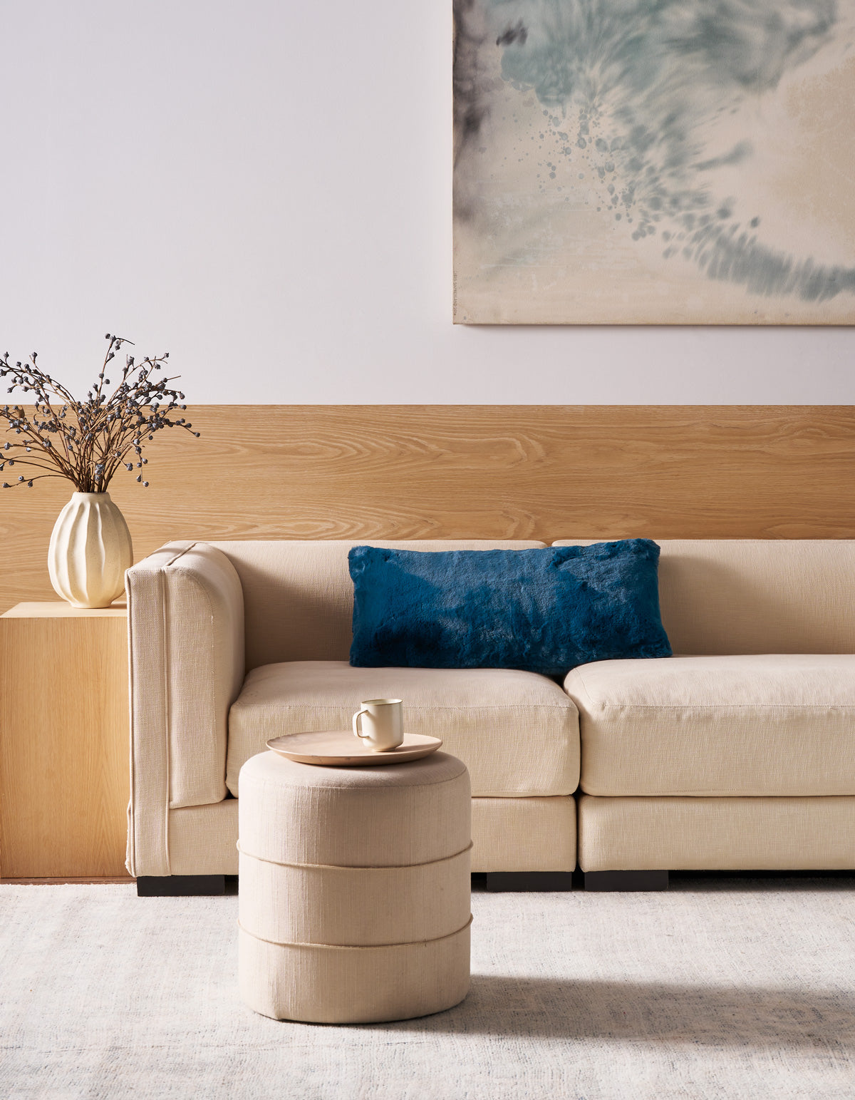A modern living room with a beige sofa, a Superplush Cuddle Pillow by Cozy Earth in blue, a round beige ottoman holding a mug, a white vase with branches, and abstract art on a wood and white wall.