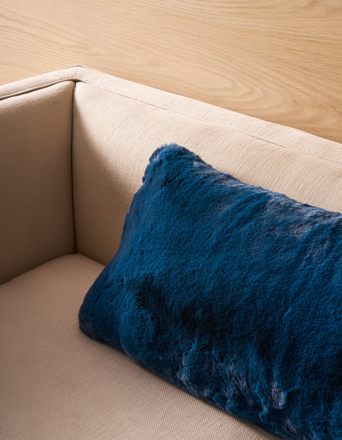 A beige fabric sofa with a Cozy Earth Superplush Cuddle Pillow in dark blue faux fur resting in the corner against a light wood-paneled wall.