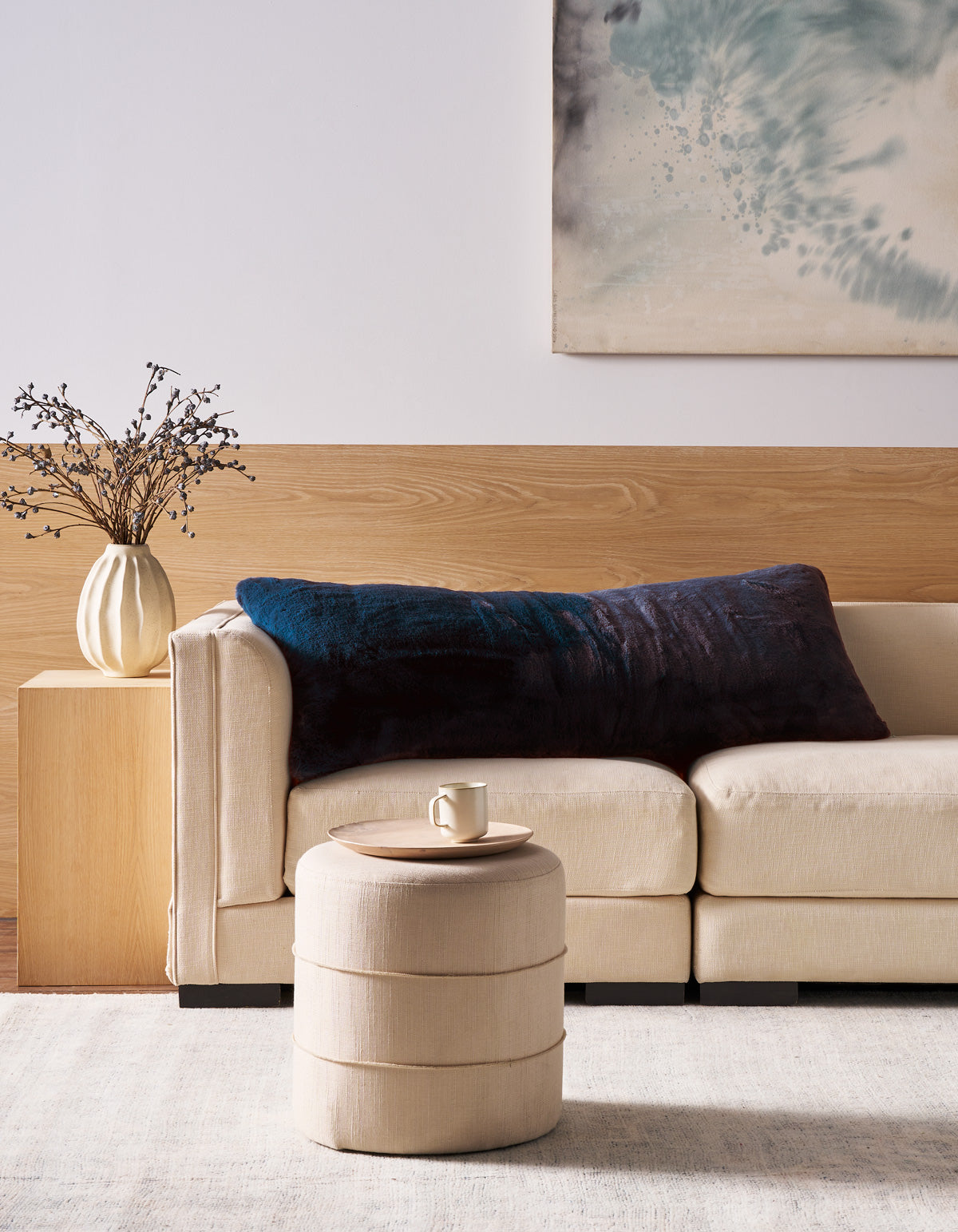 A modern living room features a beige sofa with the Superplush Cuddle Pillow by Cozy Earth in large dark blue, a round beige ottoman with a mug, a wooden side table with a vase of branches, and abstract artwork on the wall.
