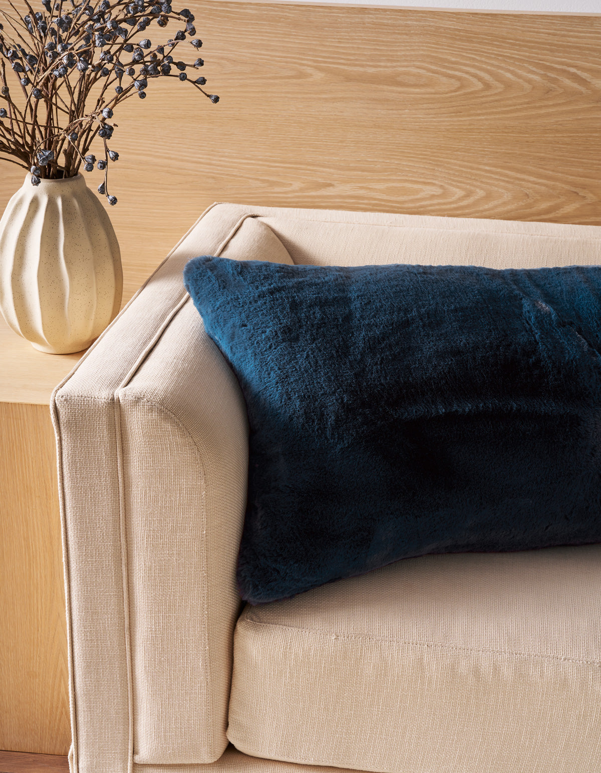 A beige sofa is adorned with the Superplush Cuddle Pillow by Cozy Earth, featuring a large, dark blue furry design. Next to the sofa, a light-colored vase holds dried branches with small round buds, set against a backdrop of light wood paneling.