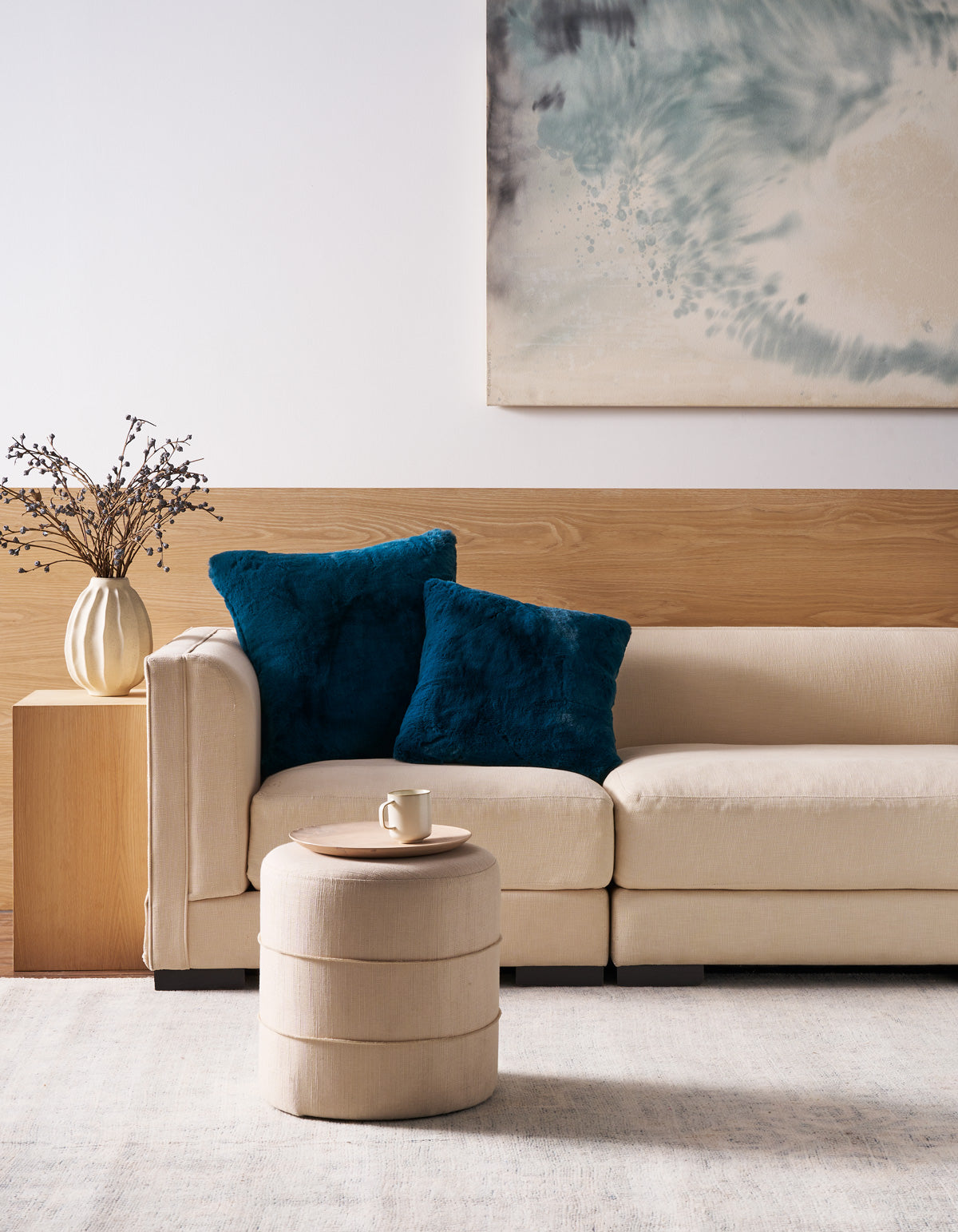 A beige sofa accented with two Cozy Earth Superplush Cuddle Pillows in blue sits against a wooden accent wall. In front, a round beige ottoman holds a cup, while the background is decorated with a vase of branches and a blue abstract painting.