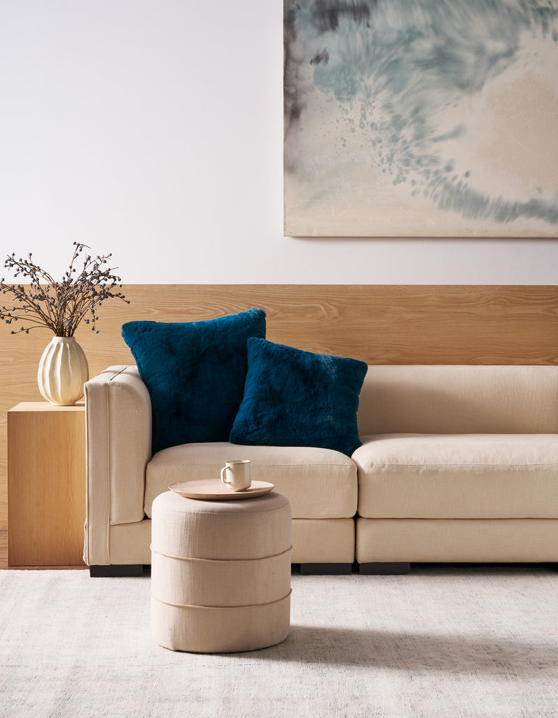 A modern living room scene featuring a beige sectional sofa with two plush dark teal throw pillows, a matching cylindrical ottoman, a wooden side table with a vase of branches, and a large abstract painting on the wall.