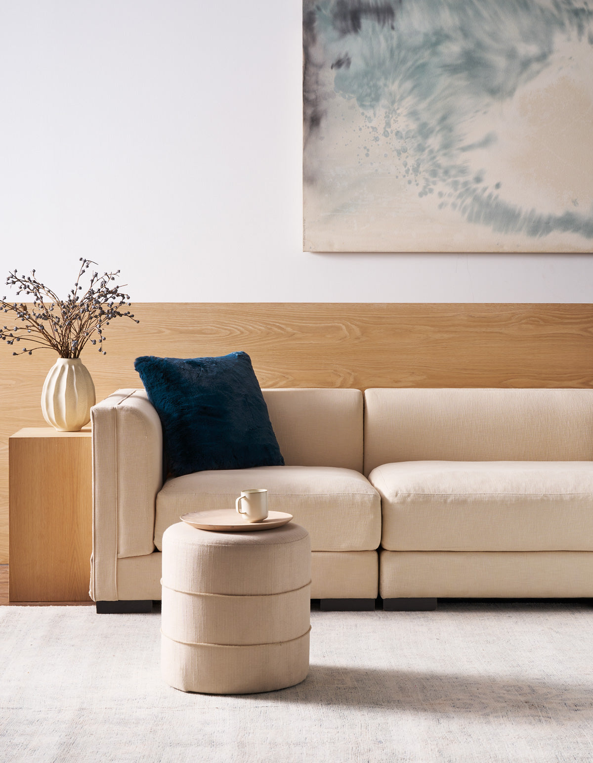 A modern living room featuring a beige sofa, the Superplush Cuddle Pillow by Cozy Earth in dark blue, a round ottoman holding a cup and saucer, a vase with dried branches, and abstract wall art in blue and gray tones displayed above wood paneling.