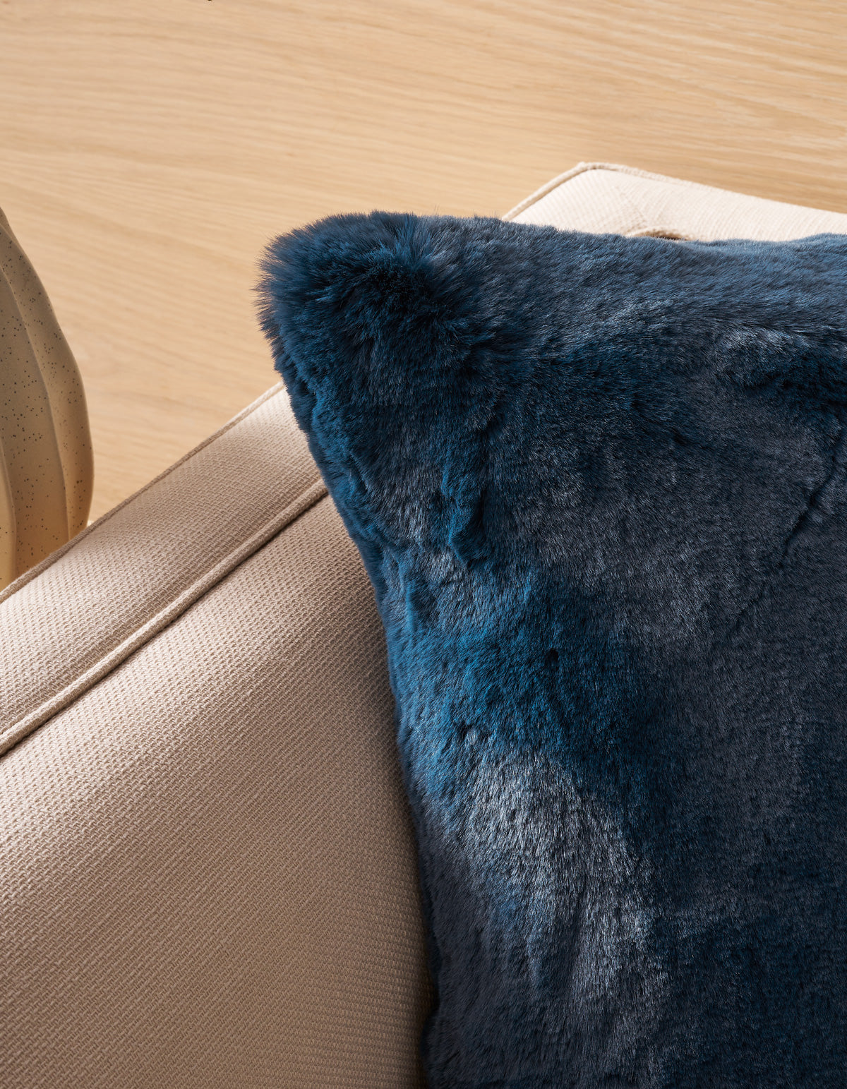 A close-up of a beige fabric couch featuring the Superplush Cuddle Pillow by Cozy Earth in dark blue faux fur, resting on the corner and set against a light wooden background.