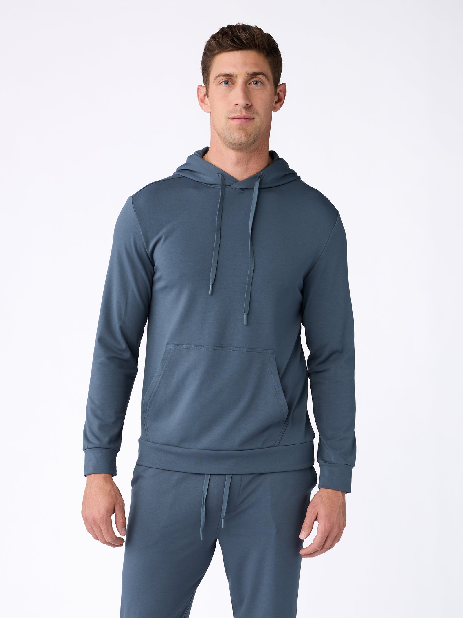 Wearing the Men's Ultra-Soft Bamboo Hoodie by Cozy Earth, a man stands against a white background in a matching blue ensemble with joggers. The pullover boasts a front pocket and drawstrings. He has short brown hair and maintains a neutral expression. 