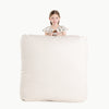 A young girl with braided hair stands behind the large Gathre Wholesale Floor Cushion in Cafe Stripe, holding the top edge and smiling at the camera against a plain white background.