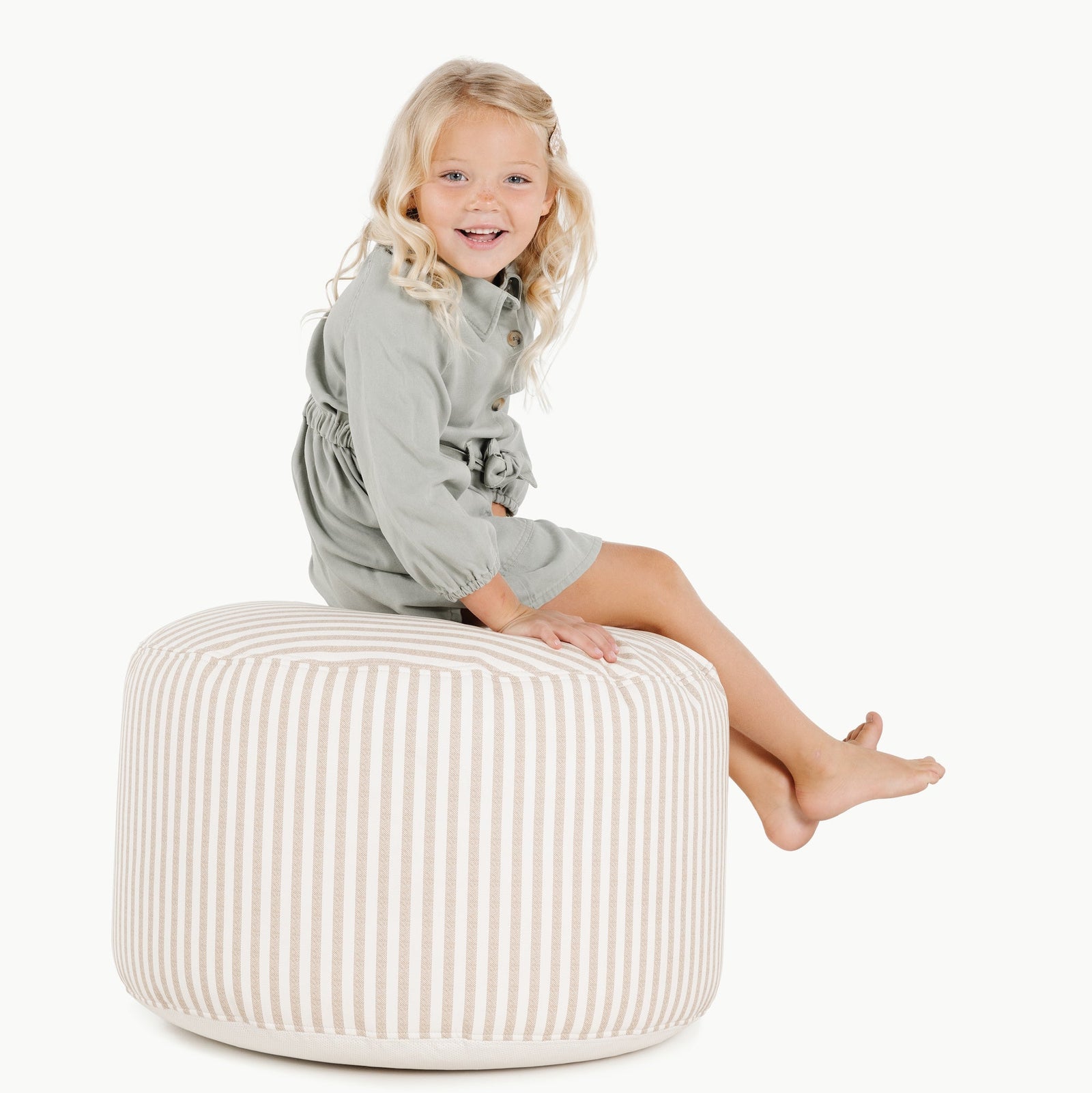 A smiling young child with blonde hair, in a light green dress, sits barefoot on the Gathre Wholesale Pouf in Cafe Stripe against a plain white background.