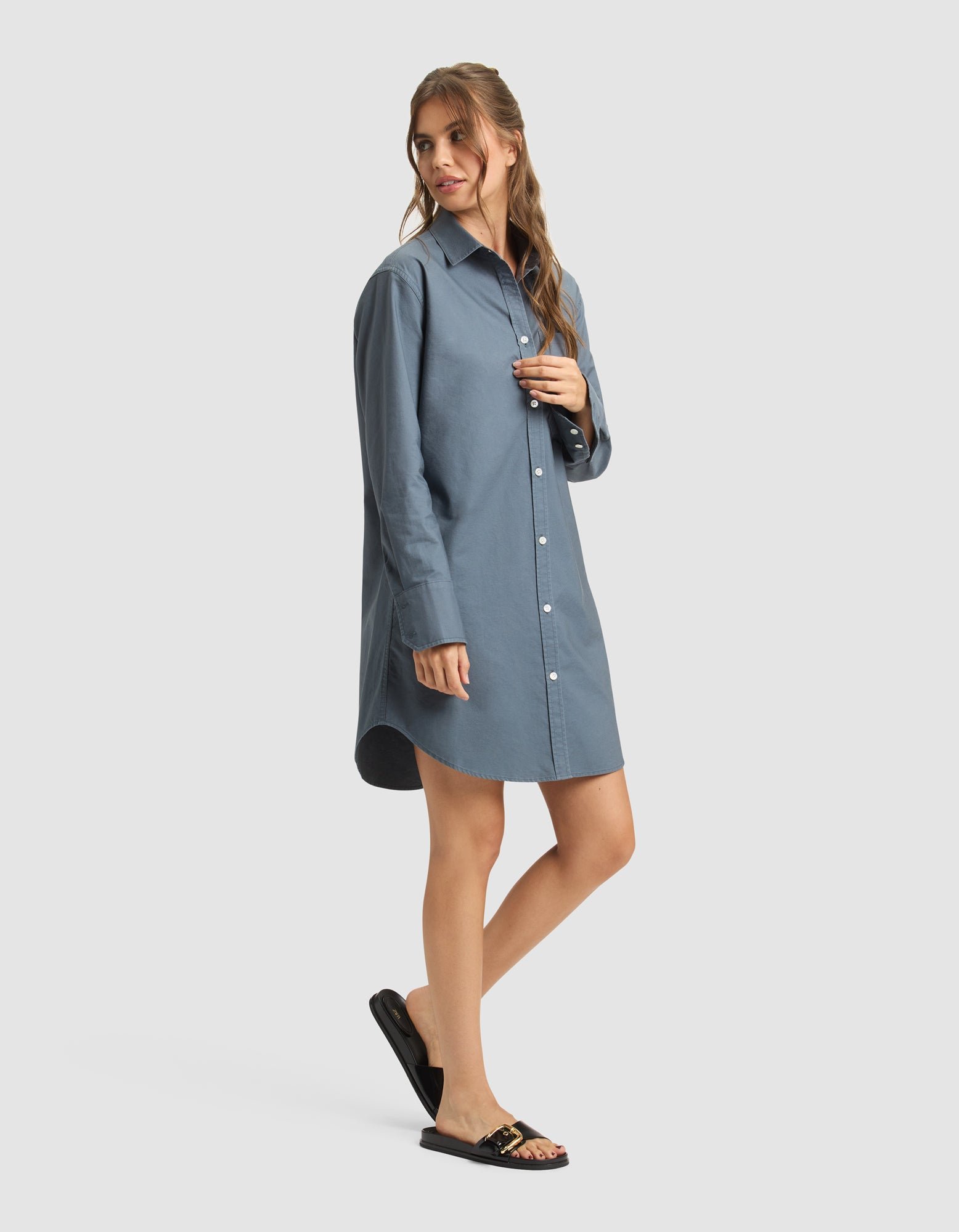 A woman poses in a relaxed way, wearing Cozy Earth's Women's Camden Dress—a loose-fitting, long-sleeve blue shirt dress—paired with black sandals featuring gold accents against a plain light background. |Color:Dawn