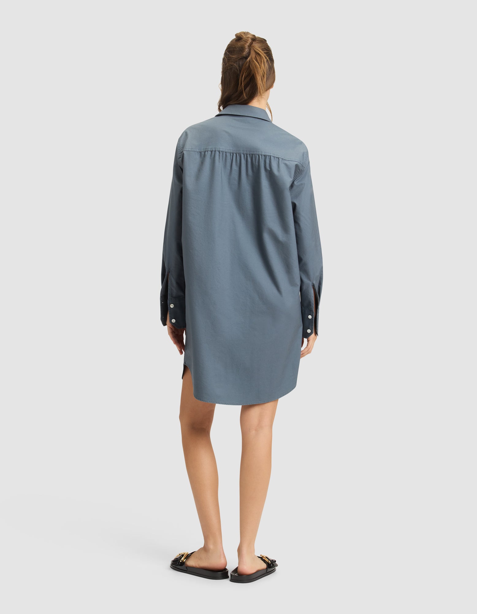 A woman with partially tied-up hair stands with her back to the camera, wearing the Cozy Earth Women's Camden Dress—a loose-fitting, long-sleeve blue shirt dress—and black flat sandals with chain details. The background is plain and light-colored. 