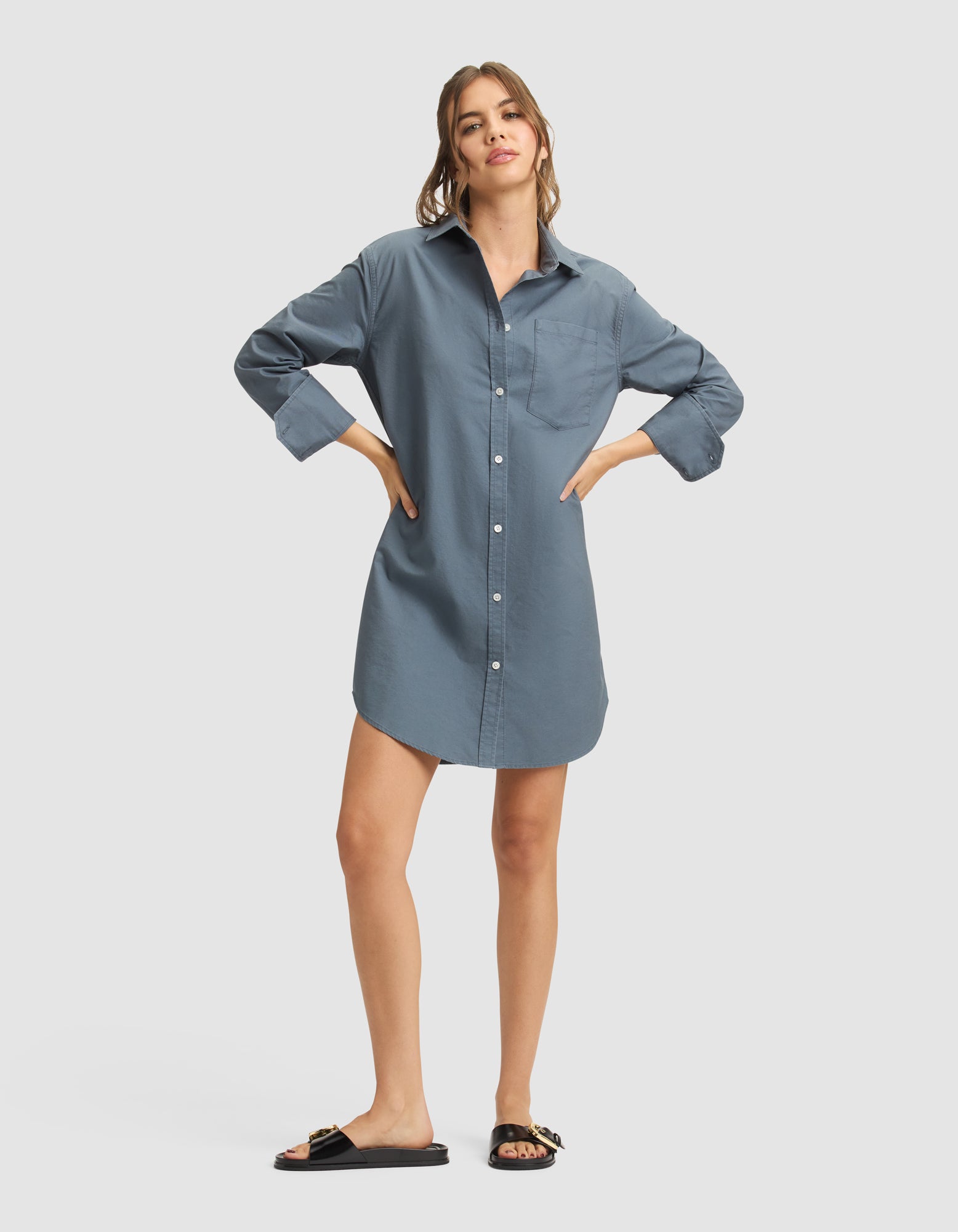 A woman wears the Cozy Earth Women's Camden Dress, a loose-fitting blue button-down shirt-dress, paired with black slide sandals with gold accents. Her long hair is pulled back and she stands against a plain light background. |Color:Dawn