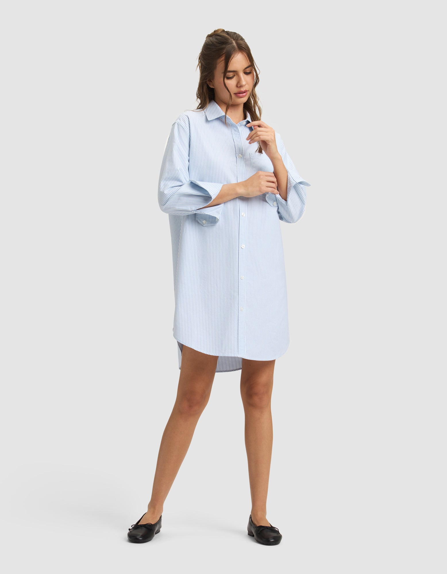 A woman stands against a plain background wearing Cozy Earth's Women's Camden Dress, an oversized light blue button-up shirt dress with long sleeves, paired with black flats. She looks down and touches her hair with one hand. 
