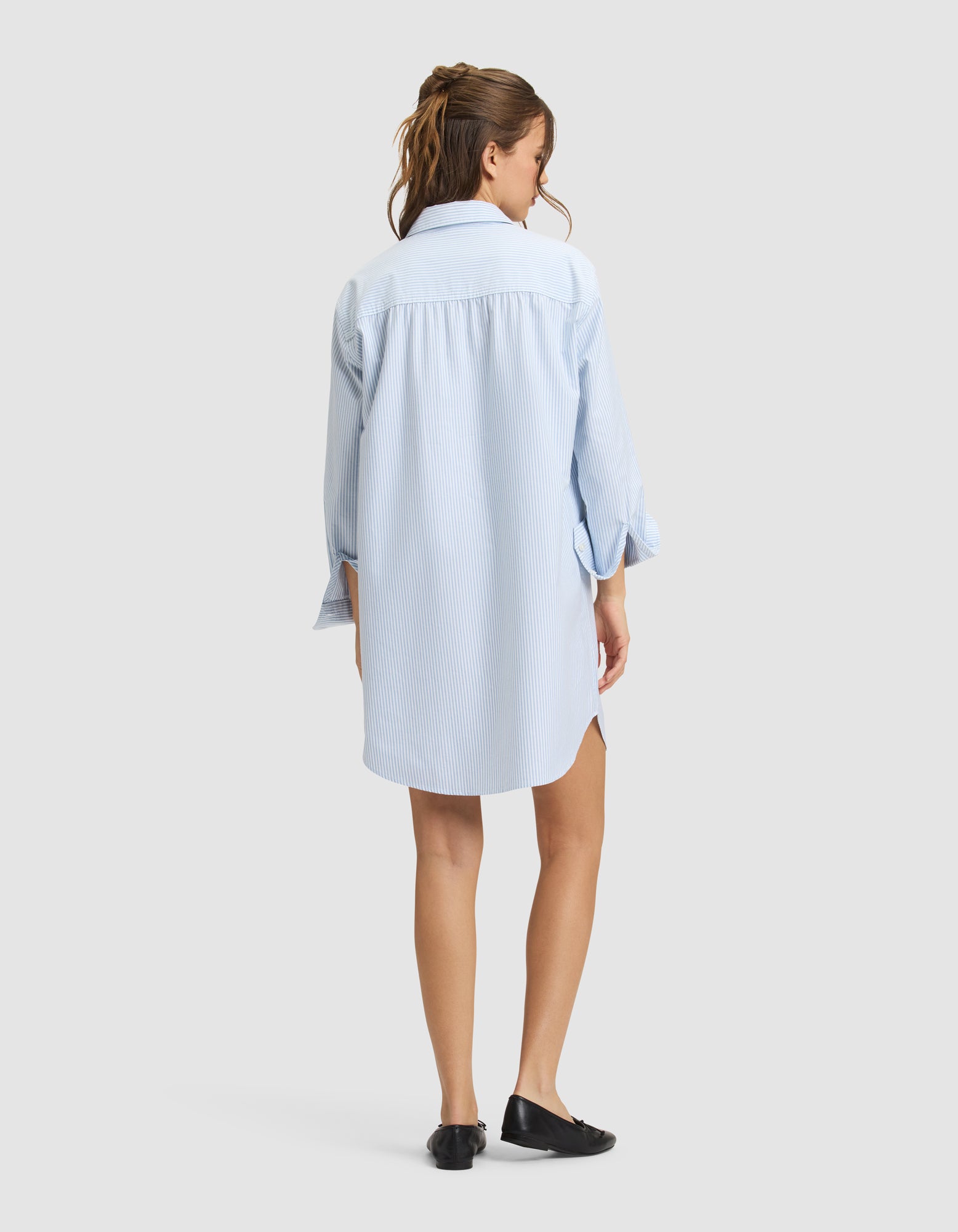A woman with long brown hair in a ponytail stands facing away, wearing the Cozy Earth Women's Camden Dress—a light blue, loose-fitting, long-sleeved shirt dress—with black shoes against a plain light gray background. 