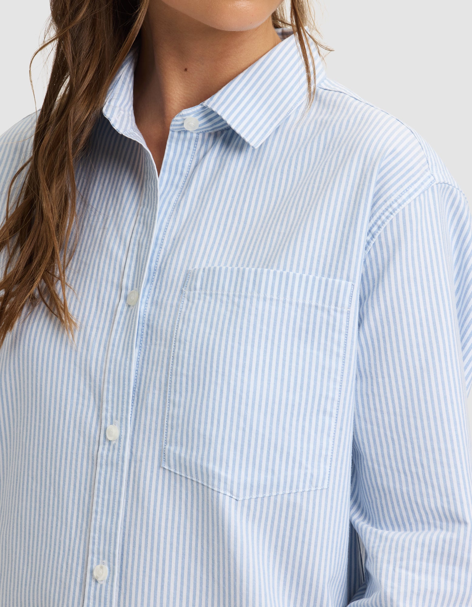 A woman wears the Cozy Earth Women's Camden Dress, featuring light blue and white vertical stripes and a chest pocket. Shown from shoulders to mid-torso, her wavy hair cascades over her shoulder. 