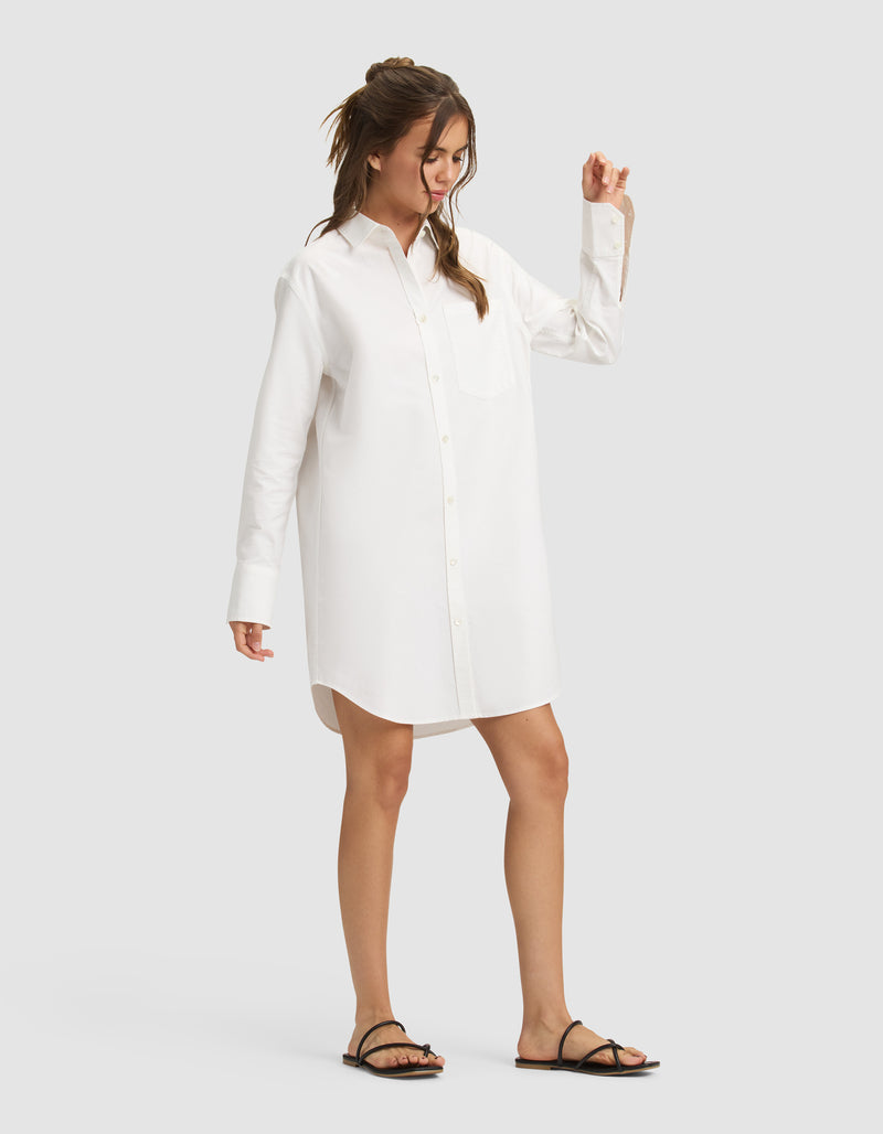 A woman models the Cozy Earth Women's Camden Dress, a loose-fitting, long-sleeve white shirt dress, with black sandals. She stands against a plain light-gray background, looking to the side with one arm slightly raised.