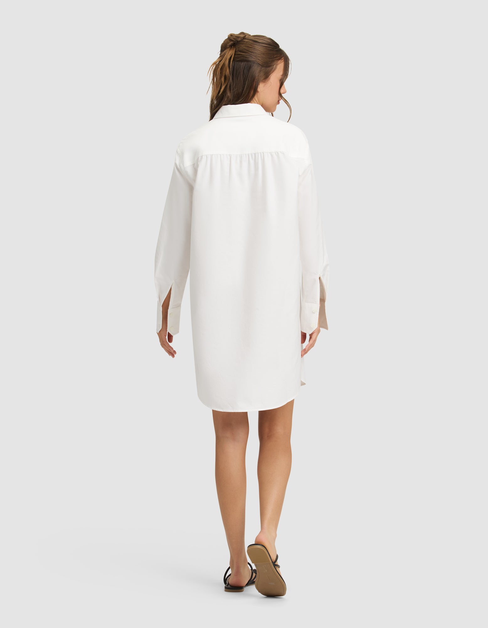 A woman with long brown hair in a loose ponytail is shown from behind, wearing the Cozy Earth Women's Camden Dress—a white, long-sleeve, knee-length shirt dress—with black sandals, standing against a plain light gray background. 