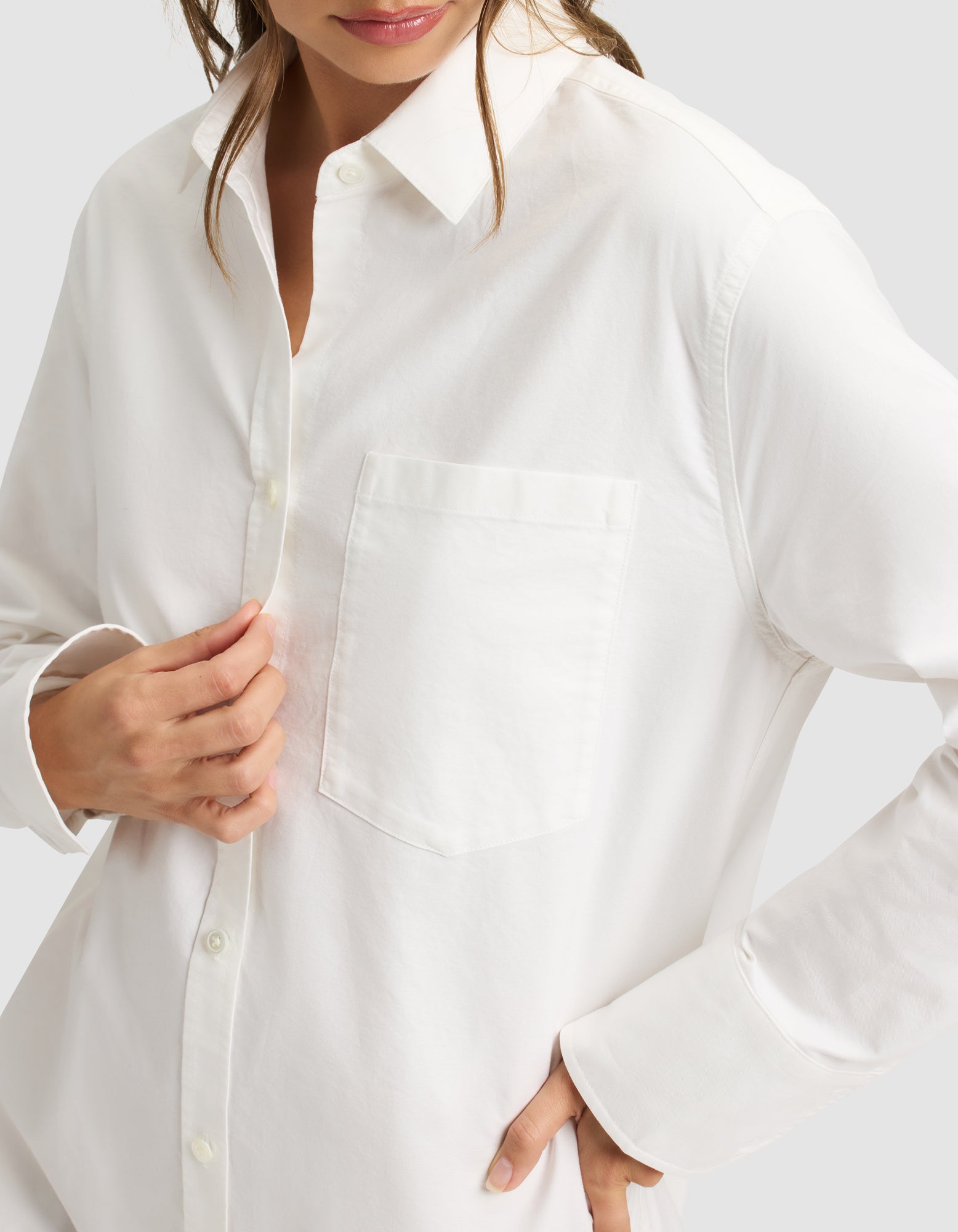 A woman is shown from shoulders to hips wearing the Cozy Earth Women's Camden Dress, a white long-sleeve button-up style with a chest pocket, partially buttoned, and touching the dress with one hand. 