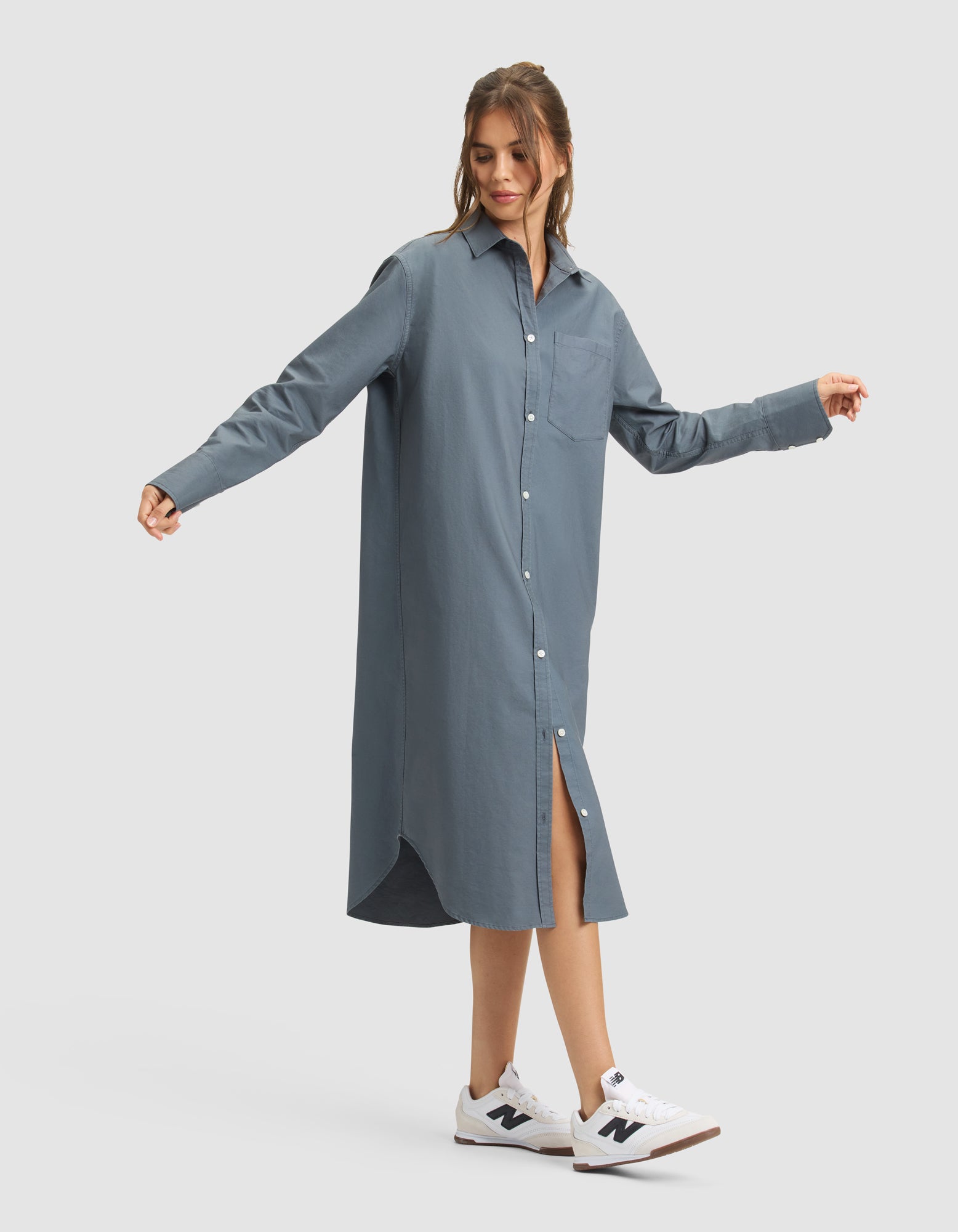 A woman models the Cozy Earth Women's Camden Midi Dress, a loose blue button-down with long sleeves and a collar, paired with white New Balance sneakers. She stands against a plain white background with arms slightly outstretched. 