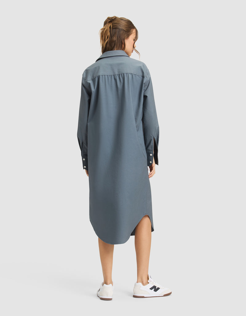 A woman faces away, wearing the Cozy Earth Women's Camden Midi Dress—a loose, long-sleeved blue button-down—paired with white sneakers on a plain light gray background.