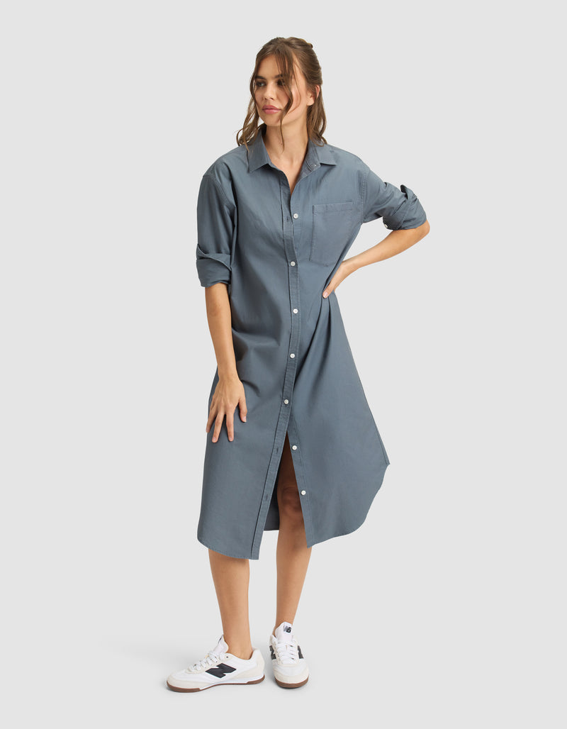 A woman stands against a light background wearing the Cozy Earth Women's Camden Midi Dress, a loose blue button-down style with rolled-up sleeves, paired with white sneakers. Her light brown hair is tied back as she poses with one hand on her hip.