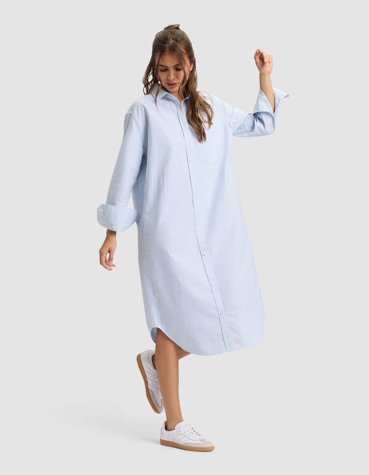 A woman wearing the Cozy Earth Women's Camden Midi Dress stands against a plain background, smiling and slightly lifting one foot. |Color:Blue Oxford Stripe