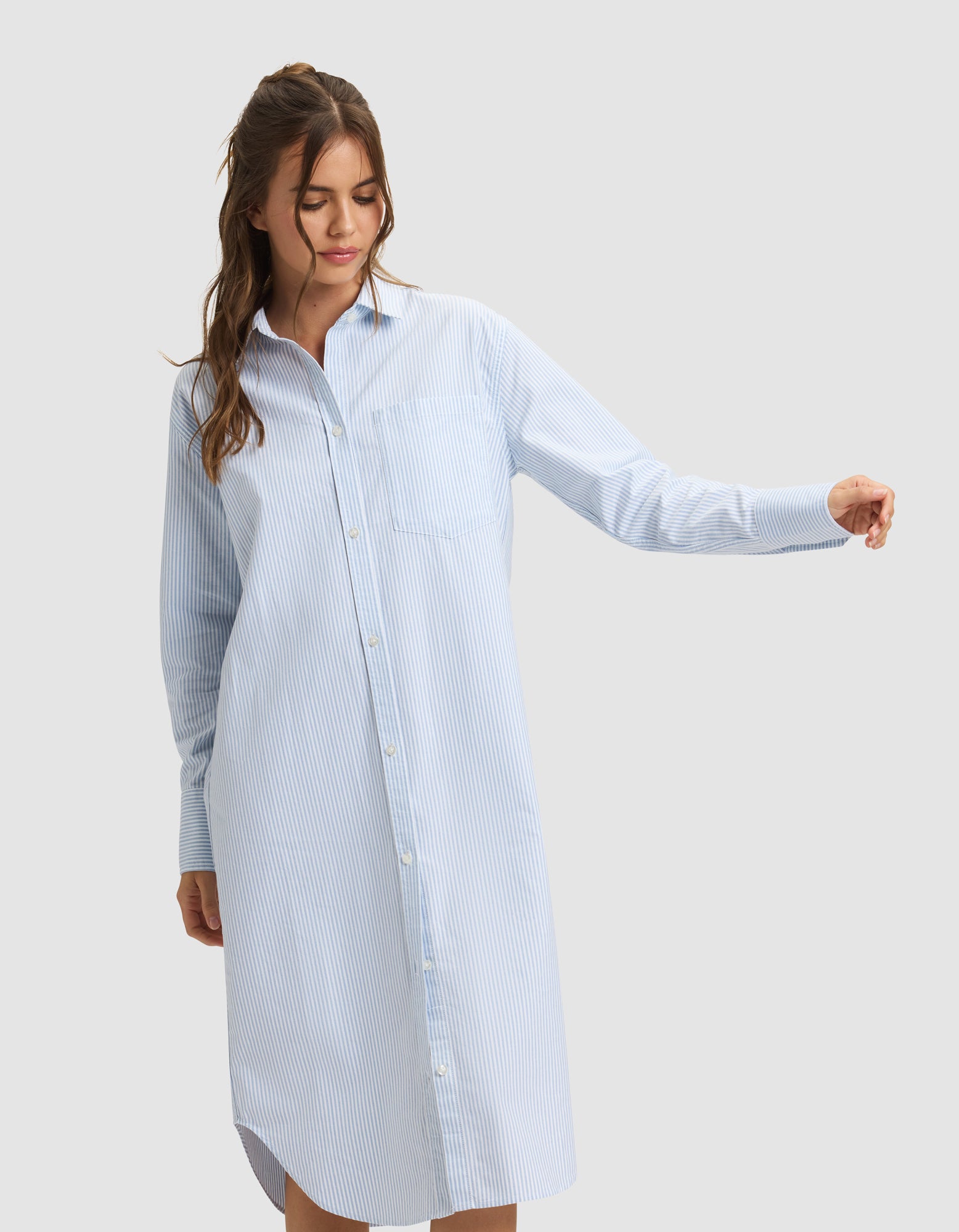 A woman in the Cozy Earth Women's Camden Midi Dress stands with one arm extended, looking down. The light blue and white striped, long-sleeve button-down dress is showcased against a plain light gray background. |Color:Blue Oxford Stripe