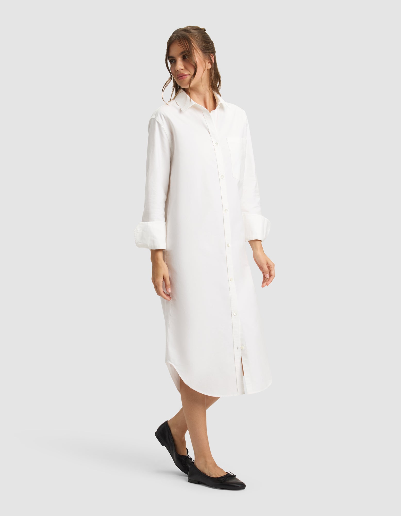 A woman models the Cozy Earth Women's Camden Midi Dress—a long white button-down shirt dress with rolled-up sleeves—paired with black flats, standing against a plain light gray backdrop. 