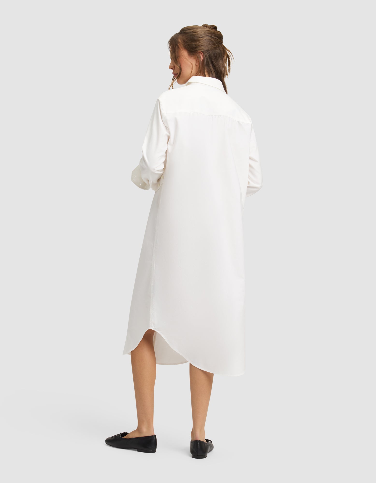 A woman with light brown hair tied up stands against a plain light gray background wearing the Cozy Earth Women's Camden Midi Dress, a white, long-sleeved, knee-length shirt dress with a curved hem, paired with black flat shoes. 