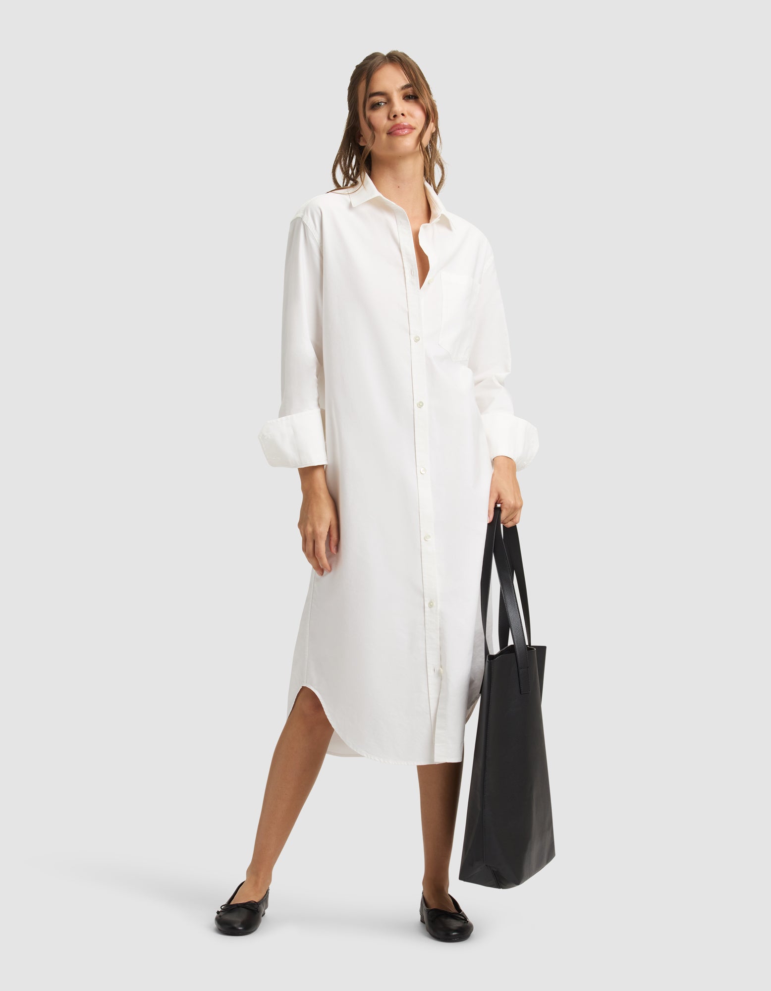 A woman smiles in a Cozy Earth Women's Camden Midi Dress, holding a large black tote bag. Her long brown hair is tied back and she wears black flats against a plain light gray background. 