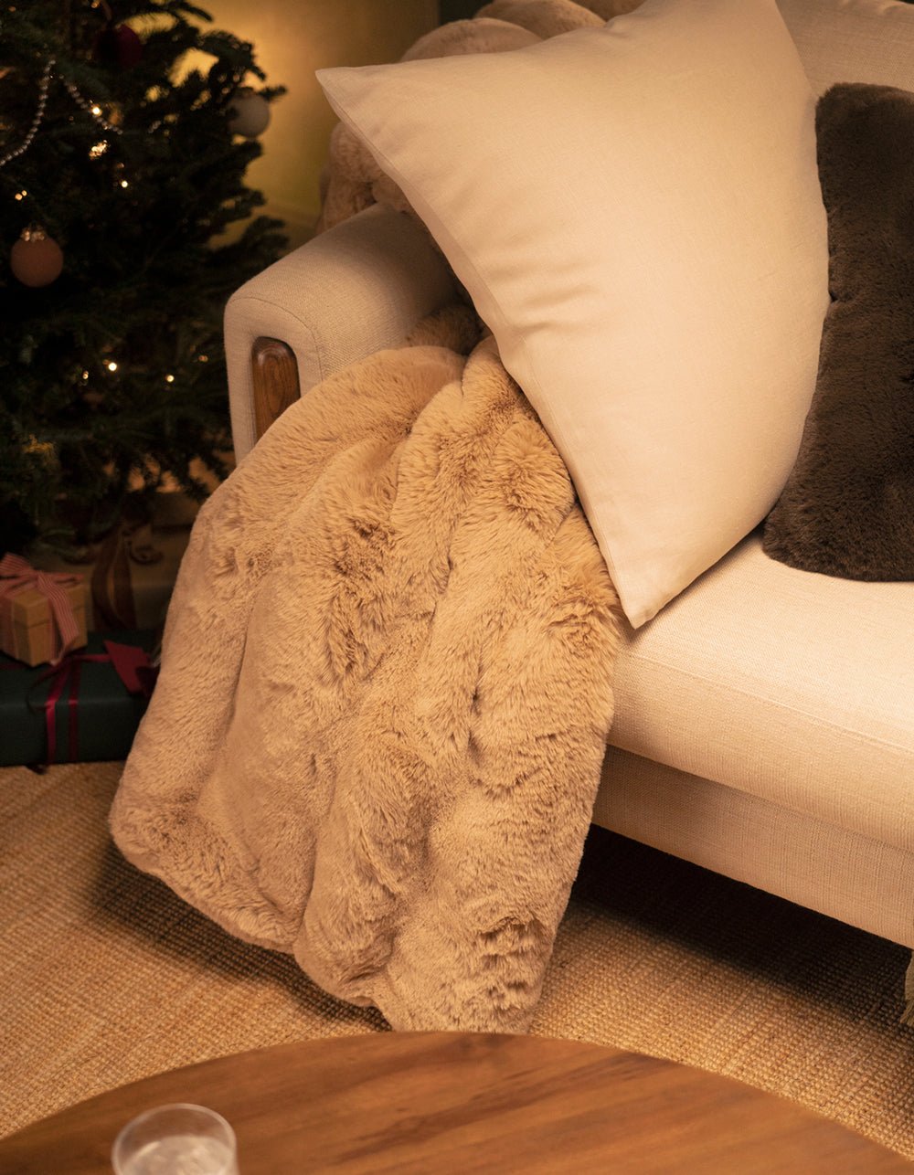 The Channel Cuddle Blanket by Cozy Earth, in beige faux fur, is draped over a cream armchair with a white pillow beside a festive Christmas tree and gifts, creating a cozy, warmly lit holiday setting.