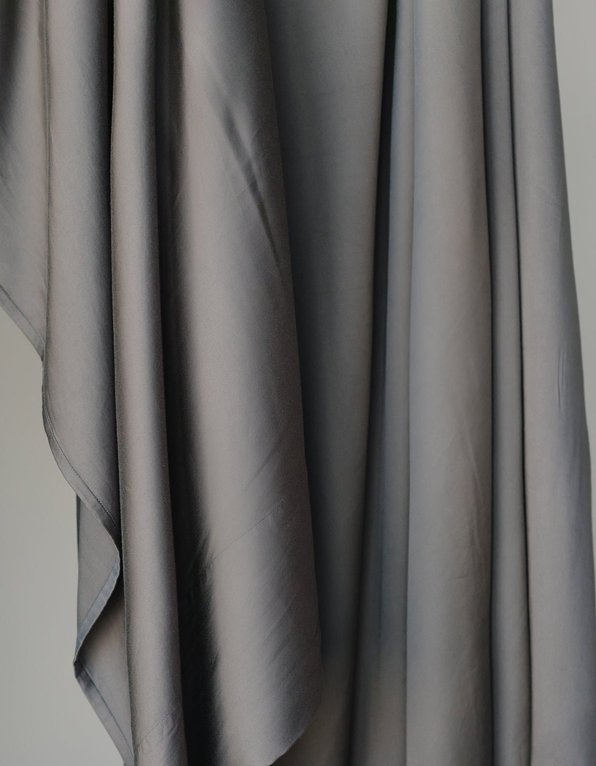 The Bamboo Sheet Set by Cozy Earth features smooth, draped gray fabric with gentle folds and a soft texture, creating elegant waves and shadows as it cascades down. 