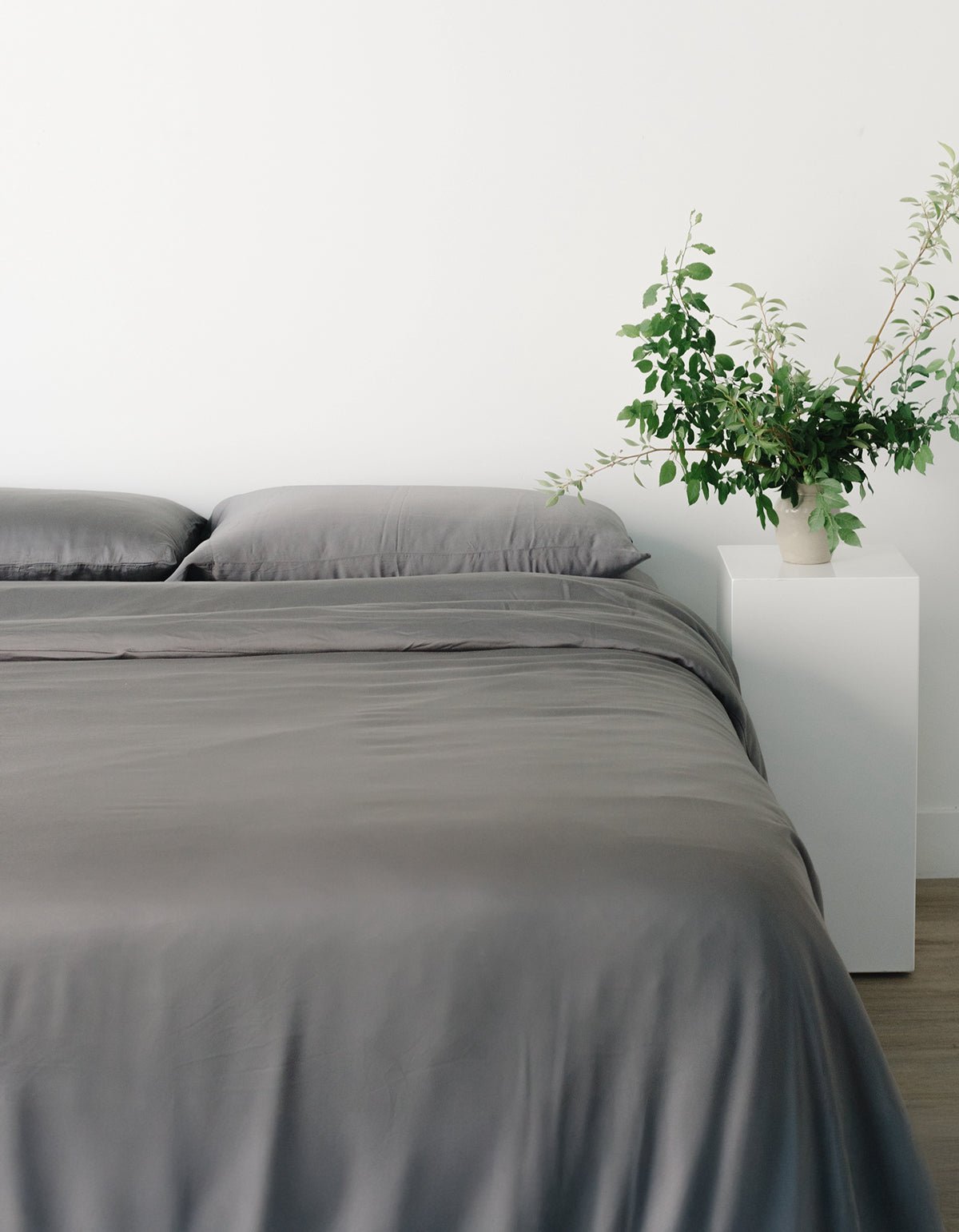 A neatly made bed with Cozy Earth's Bamboo Sheet Set in gray and two pillows sits beside a white nightstand holding a potted plant with green foliage. The minimalist room features a simple white wall background. 