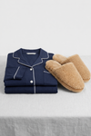 The Wind-Down Bundle from Cozy Earth, featuring navy blue pajamas with white piping and tan fuzzy slippers, rests on a light gray fabric surface.