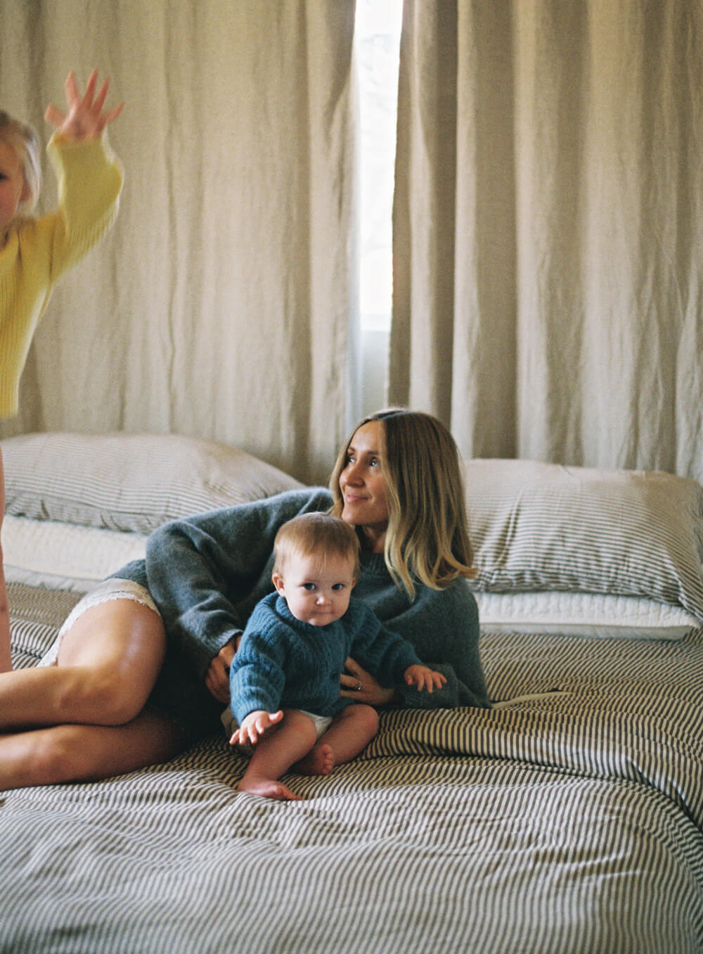 Woman lying on a bed with a baby and another child in a room with neutral-colored curtains.