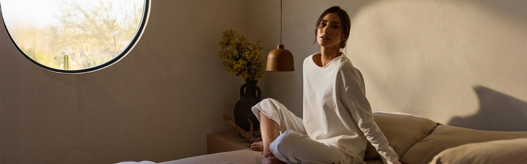 A woman wearing Cozy Earth Chelsea Set sits in a room with a large, round window. She sits on a bed next to a vase of flowers and pendulum lamp.