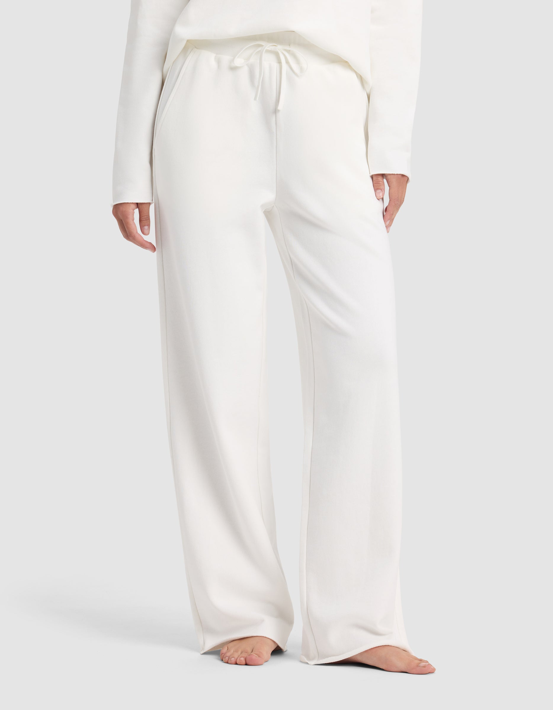 A person is shown barefoot from shoulders to feet against a plain light background, wearing Cozy Earth's Chelsea Pant—a pair of loose-fitting, white drawstring lounge pants—with a matching long-sleeve top.