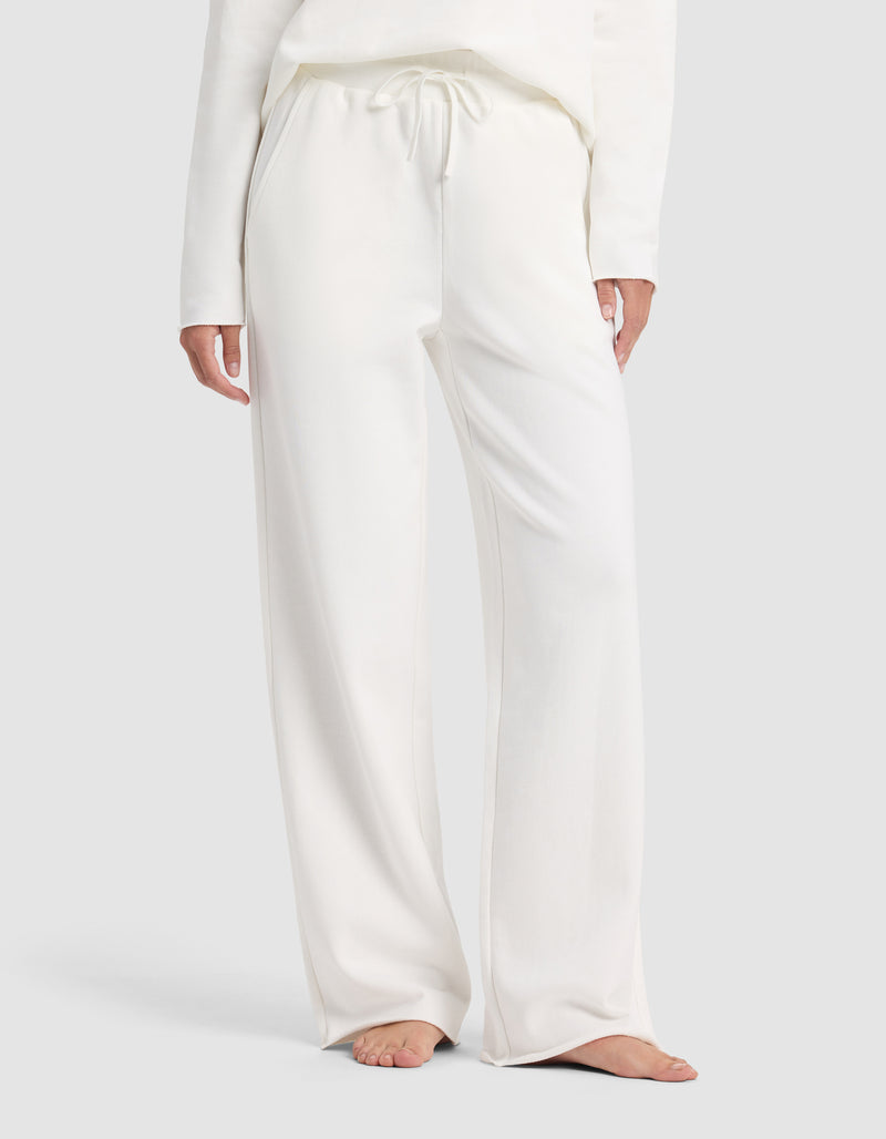 A person is shown barefoot from shoulders to feet against a plain light background, wearing Cozy Earth's Chelsea Pant—a pair of loose-fitting, white drawstring lounge pants—with a matching long-sleeve top.