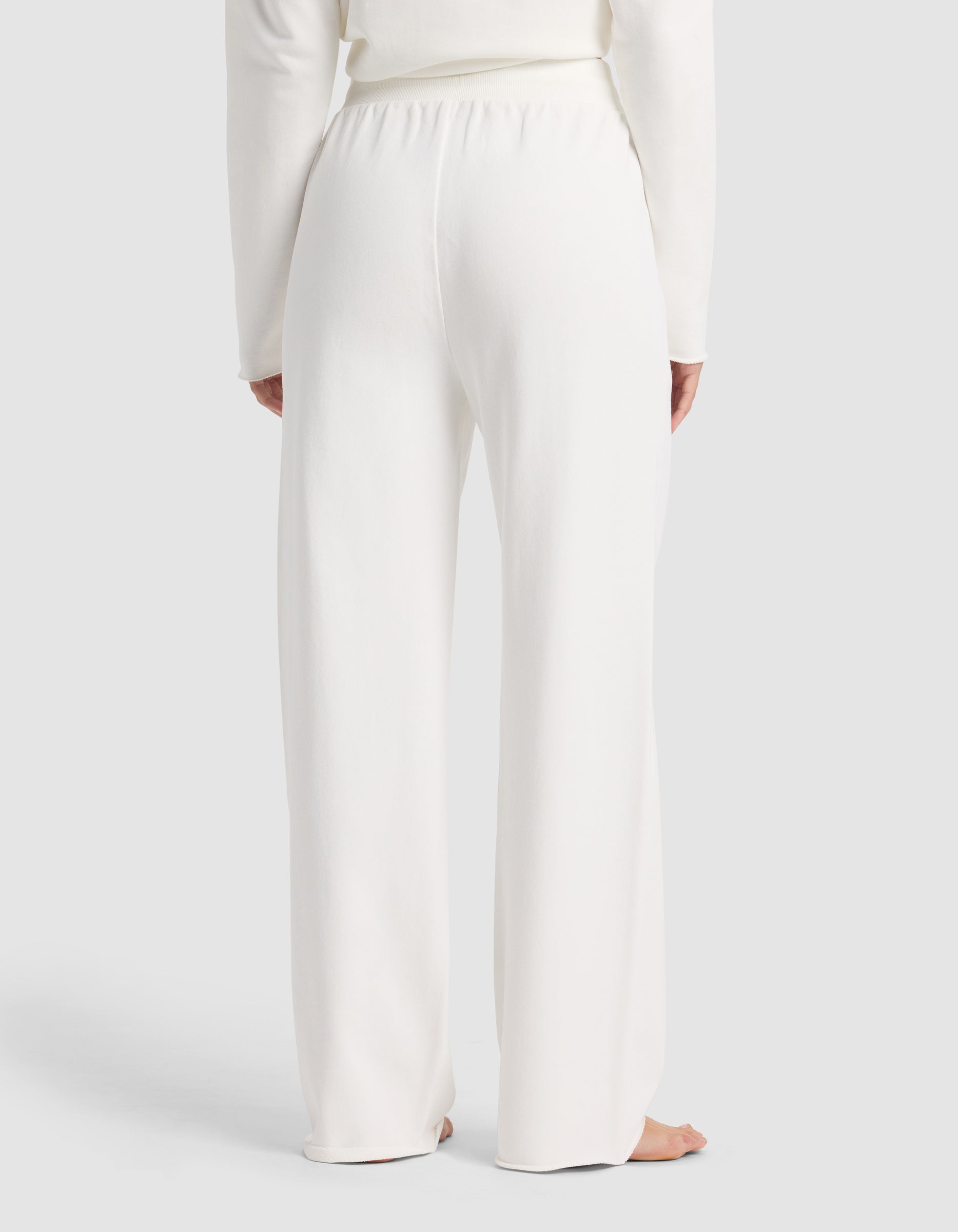 A person stands barefoot facing away, wearing Cozy Earth's Chelsea Pant—loose-fitting, high-waisted white lounge pants—with a matching long-sleeve top against a plain light background.|Color:Bone