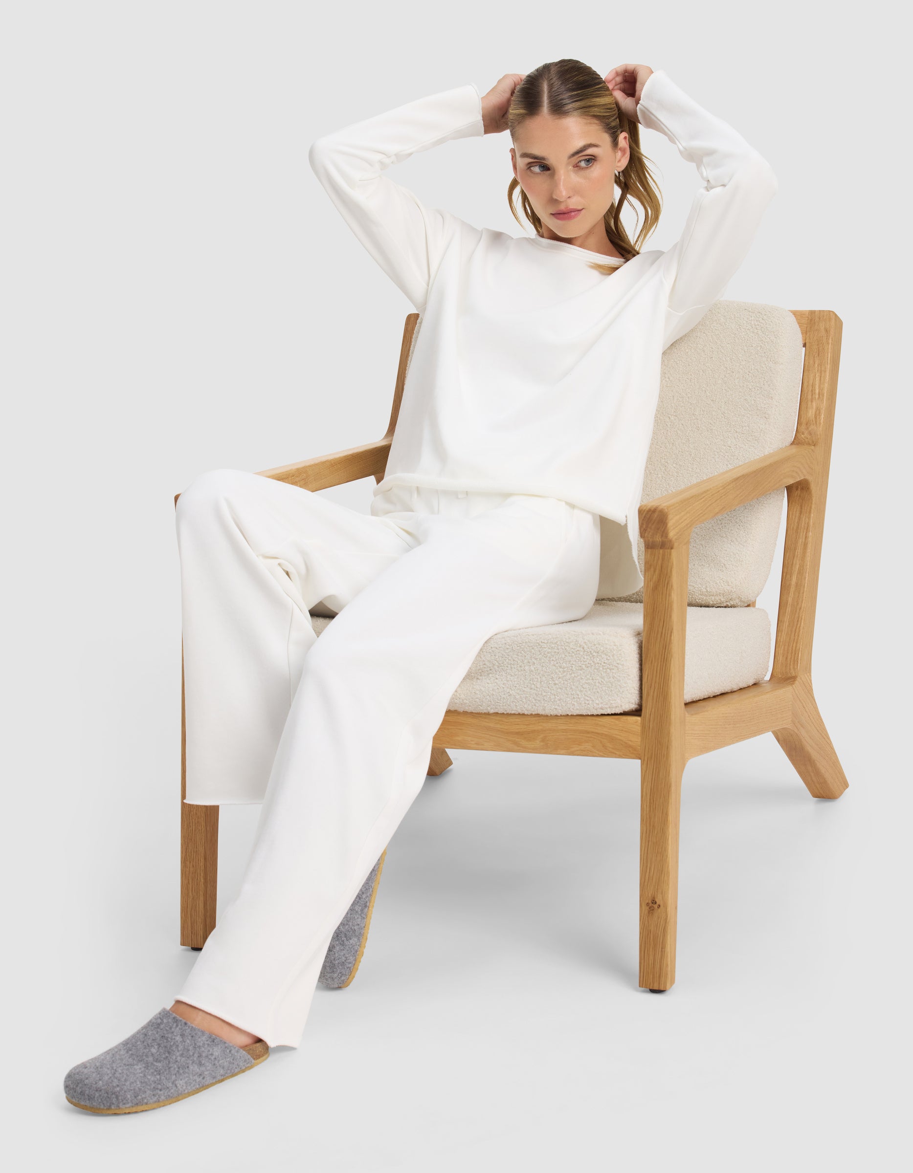 A woman wearing Cozy Earth's Chelsea Pant and gray slippers sits on a wooden armchair with cream cushions, adjusting her hair and looking to the side against a plain, light background.|Color:Bone