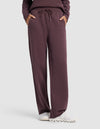 Model wearing Cozy Earth Chelsea Pant in dark maroon with hands in pockets, paired with a matching top and white sneakers, standing against a plain light background.|Color:Fig